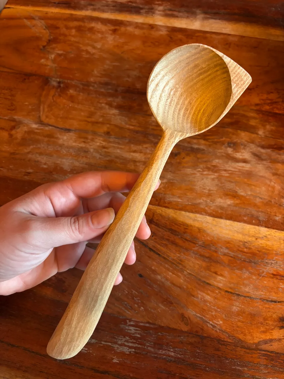 A pale catalpa spoon with a pointed corner on the bowl, held over a wooden table
