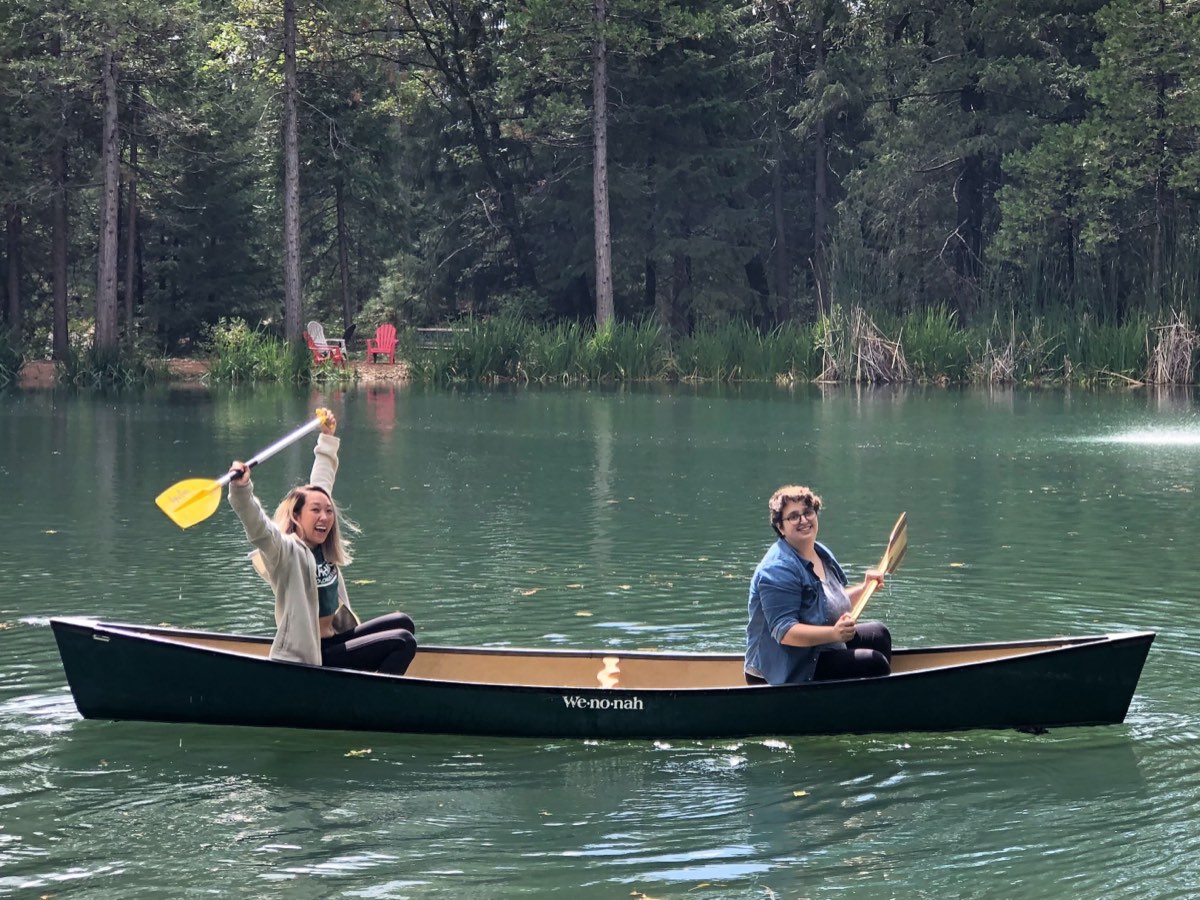 Maggie and Anna sitting at either end of a dark green canoe on a lake ringed with pine trees, both smiling, one raising a yellow paddle over her head